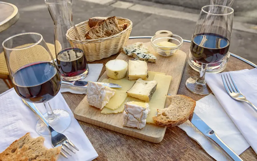 A pavement cafe table in Paris set with two glasses and a carafe of red wine, a wooden board of French cheeses, a basket of sliced baguette and white napkins