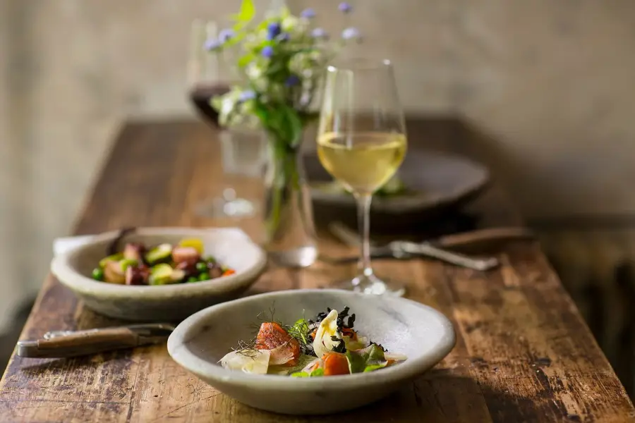 A rustic wooden table laid with two plated dishes, a glass of white wine in the foreground and a glass of red wine behind