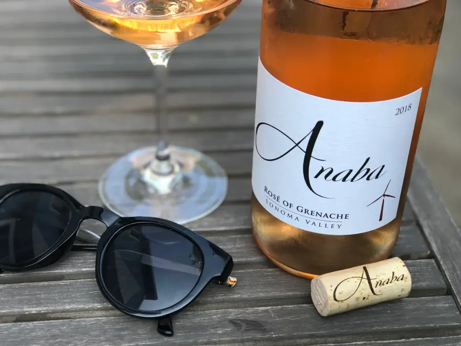 A glass of pale rosé wine glowing in low evening sun on a weathered wooden table, next to a bottle and a pair of sunglasses