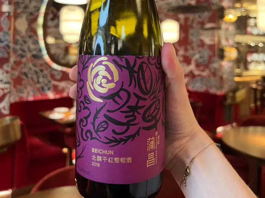 A bottle of Puchang Beichun 2018 held up close in a red-walled restaurant, its magenta label showing a gold rose, black calligraphic flourishes and the words BEICHUN 北醇干红葡萄酒 2018
