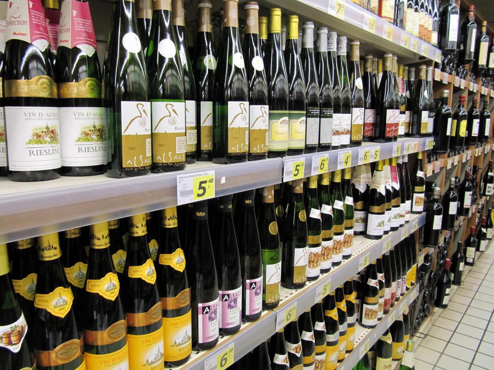 A supermarket shelf lined with tall green bottles of Alsace wine, price labels on the rail beneath each row