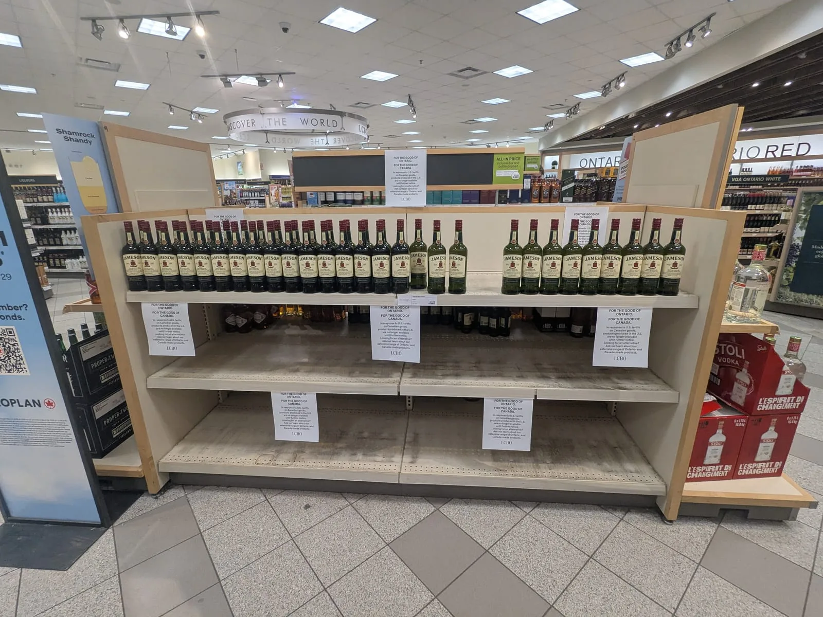 Bare wooden shelving in an Ontario liquor store where American wine and spirits used to be stocked, with empty price rails and a few remaining bottles at the far end