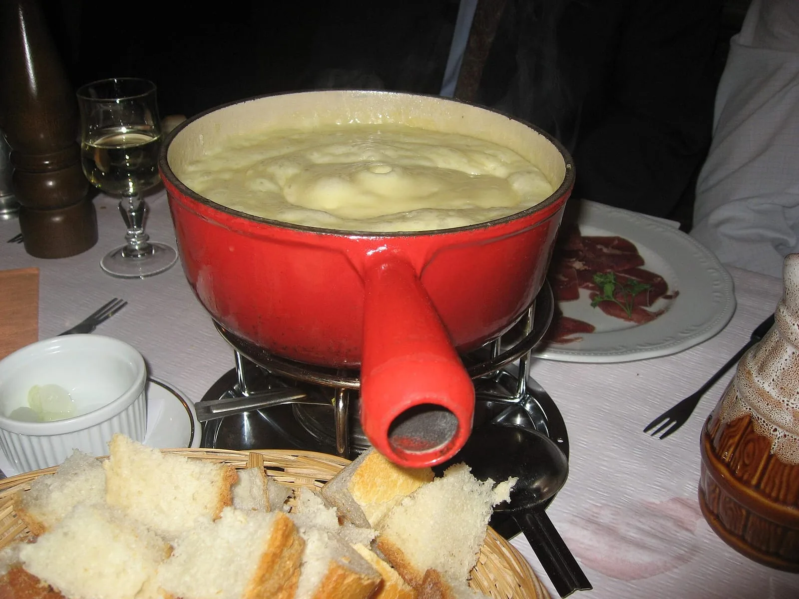 A red caquelon of cheese fondue on its burner with bread cubes and a glass of white wine