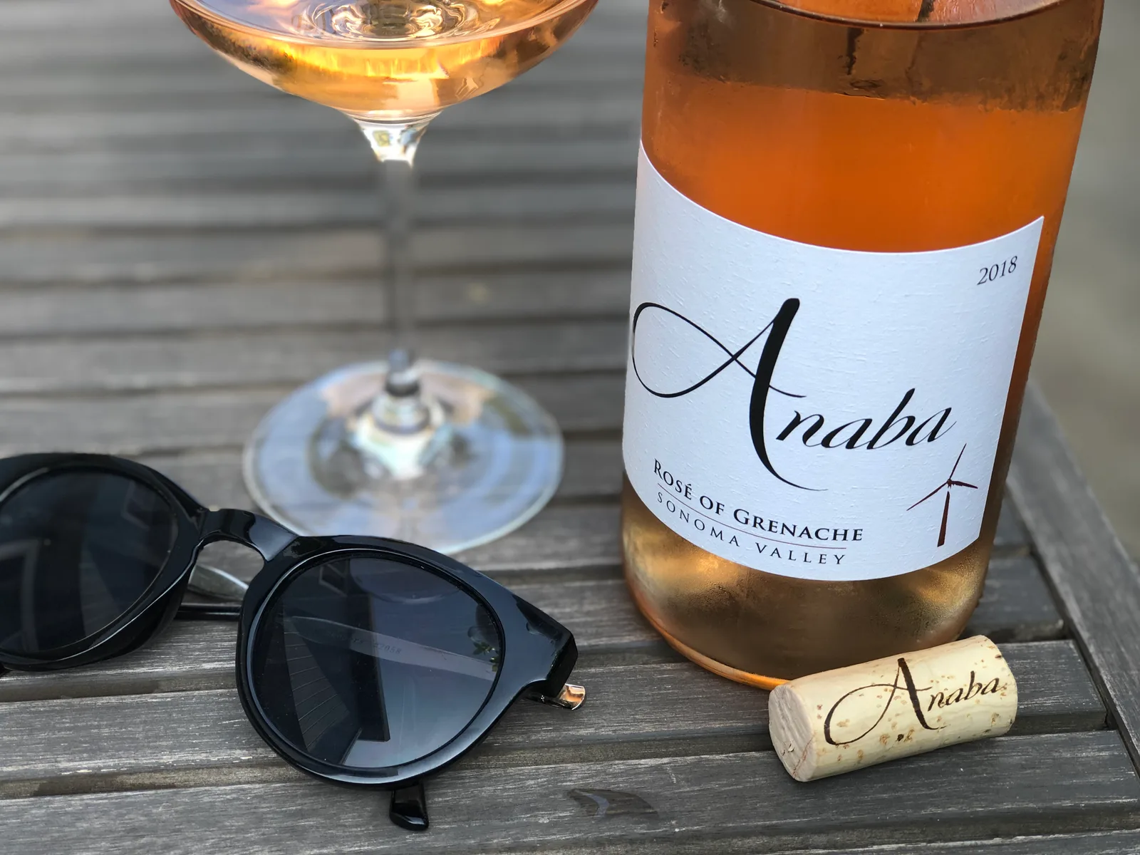 A glass of pale rosé wine glowing in low evening sun on a weathered wooden table, next to a bottle and a pair of sunglasses