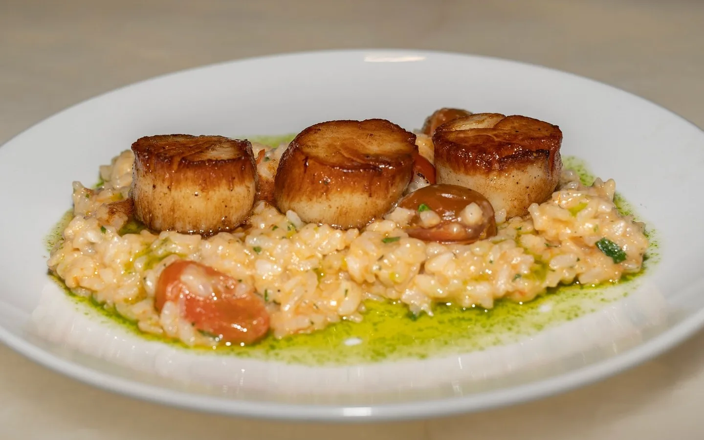 Three pan-seared scallops with a deep golden crust sitting on creamy tomato risotto finished with herb oil, on a white plate