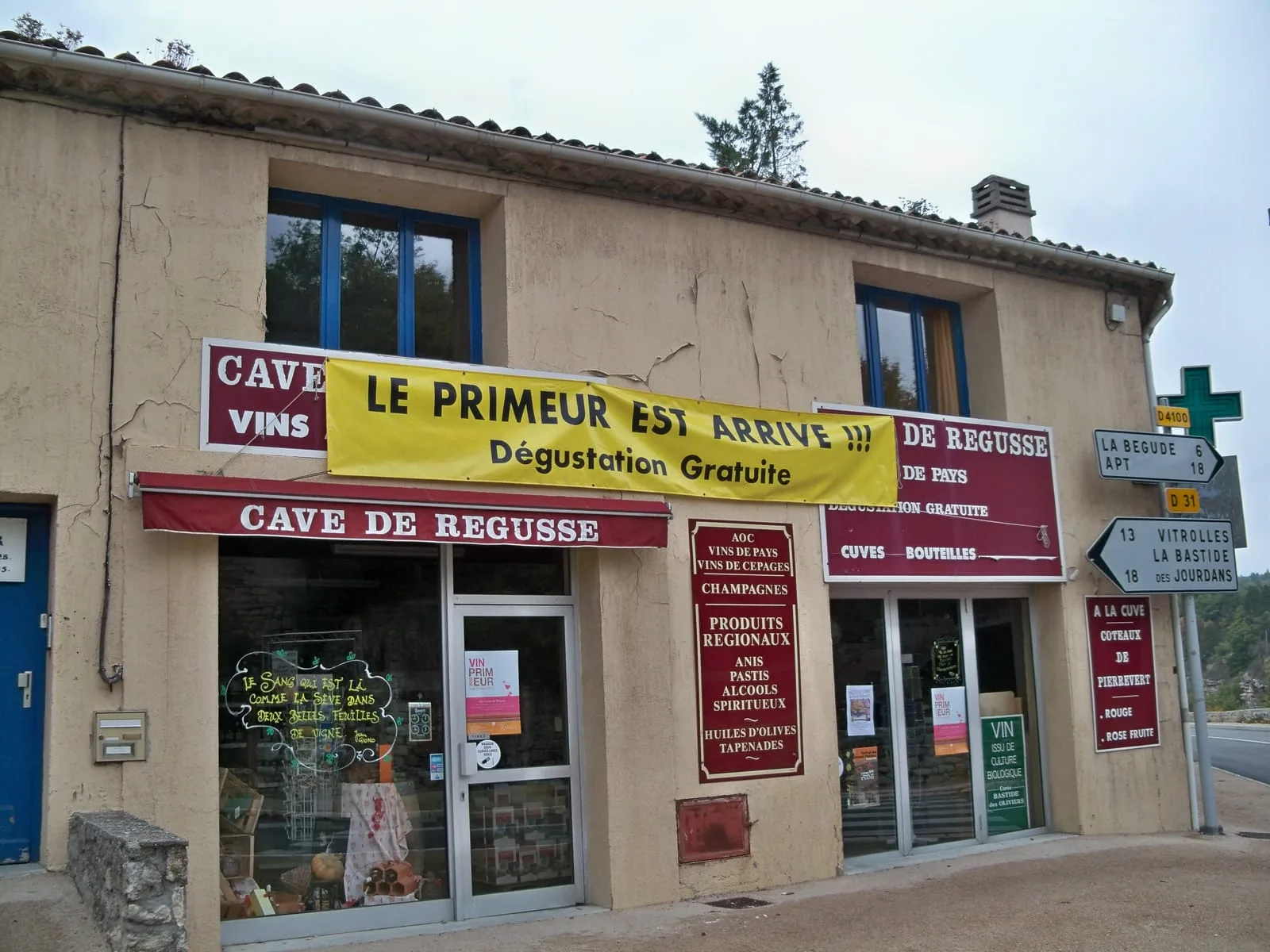 The front of a small village wine shop in Provence, its offer hand-painted across red panels and a yellow banner, with road signs beside it