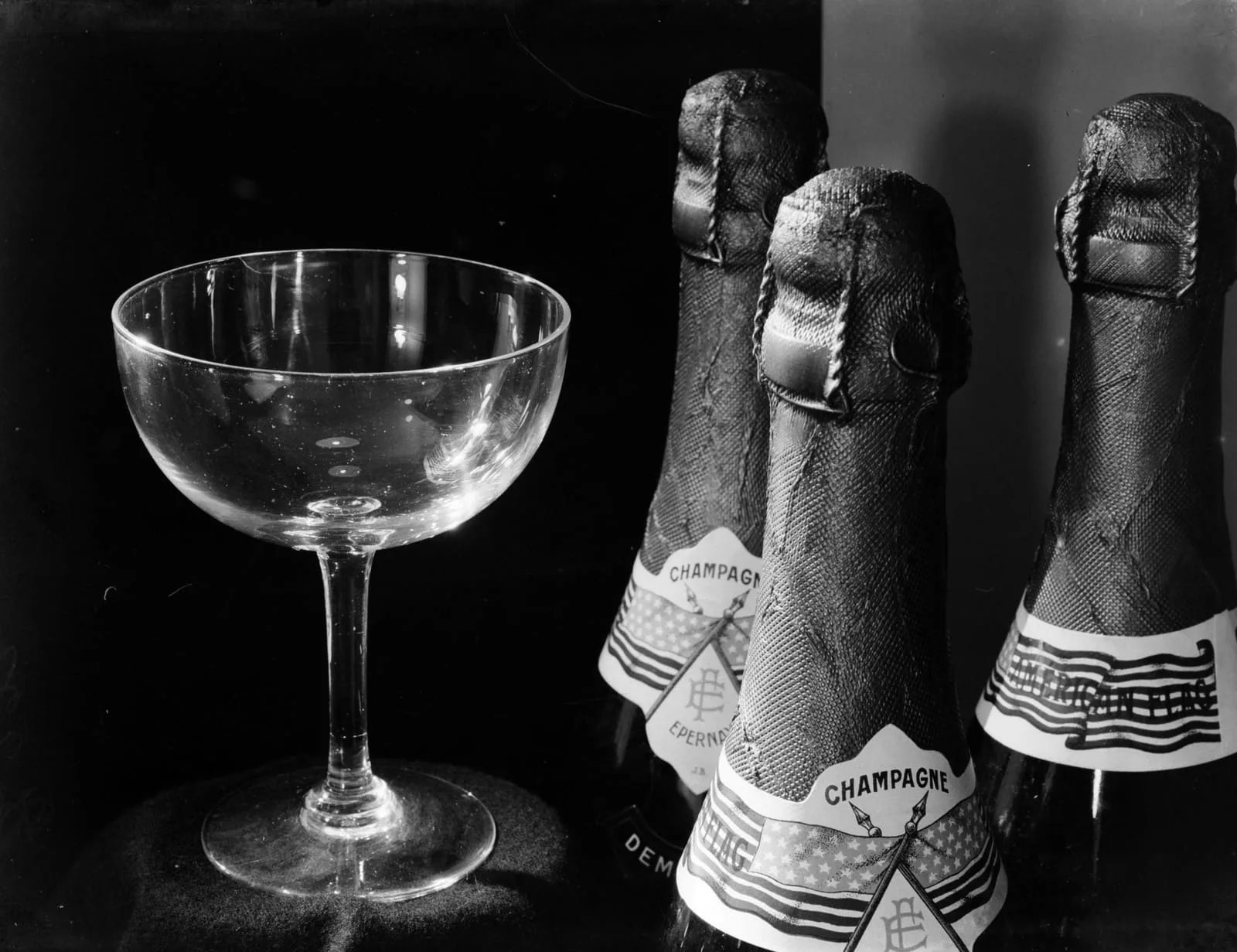 Three Champagne bottles with foil-wrapped necks and Épernay labels standing beside an empty coupe glass, photographed in black and white against a dark background