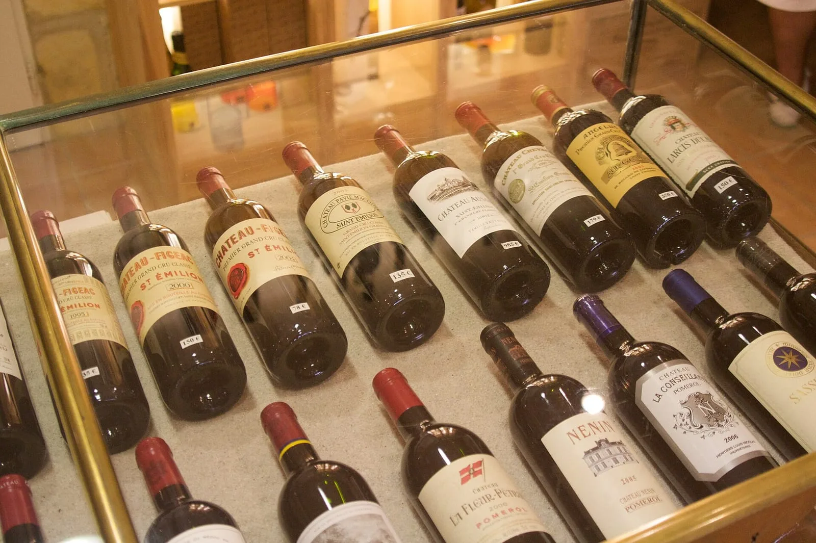 Bottles of Saint-Émilion and Pomerol laid on their sides in a locked glass retail display case, each with a small white price tag propped against it