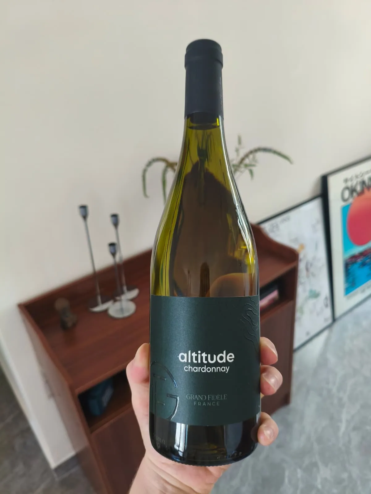 The full bottle of Grand Fidèle Altitude Chardonnay, dark green label and black capsule, held in front of a living room sideboard