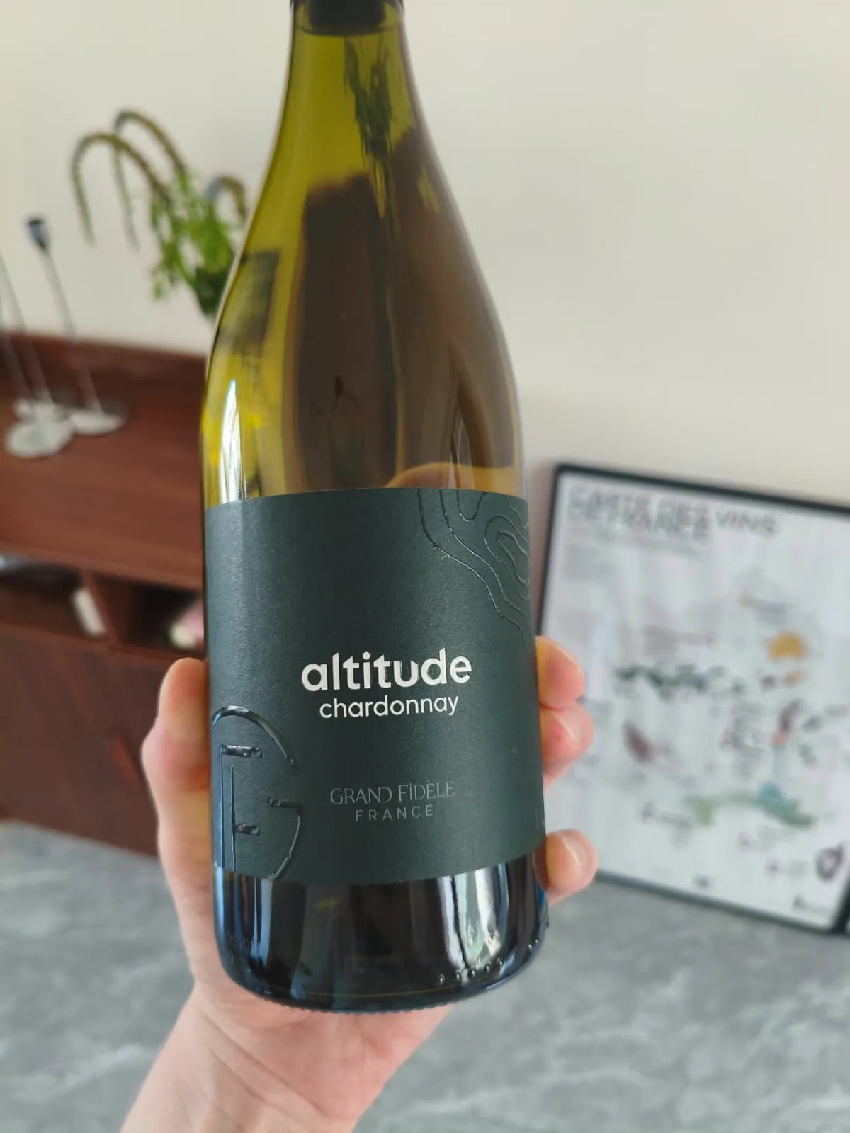 A bottle of Grand Fidèle Altitude Chardonnay held up close, its dark green label showing embossed contour lines and the estate's G monogram