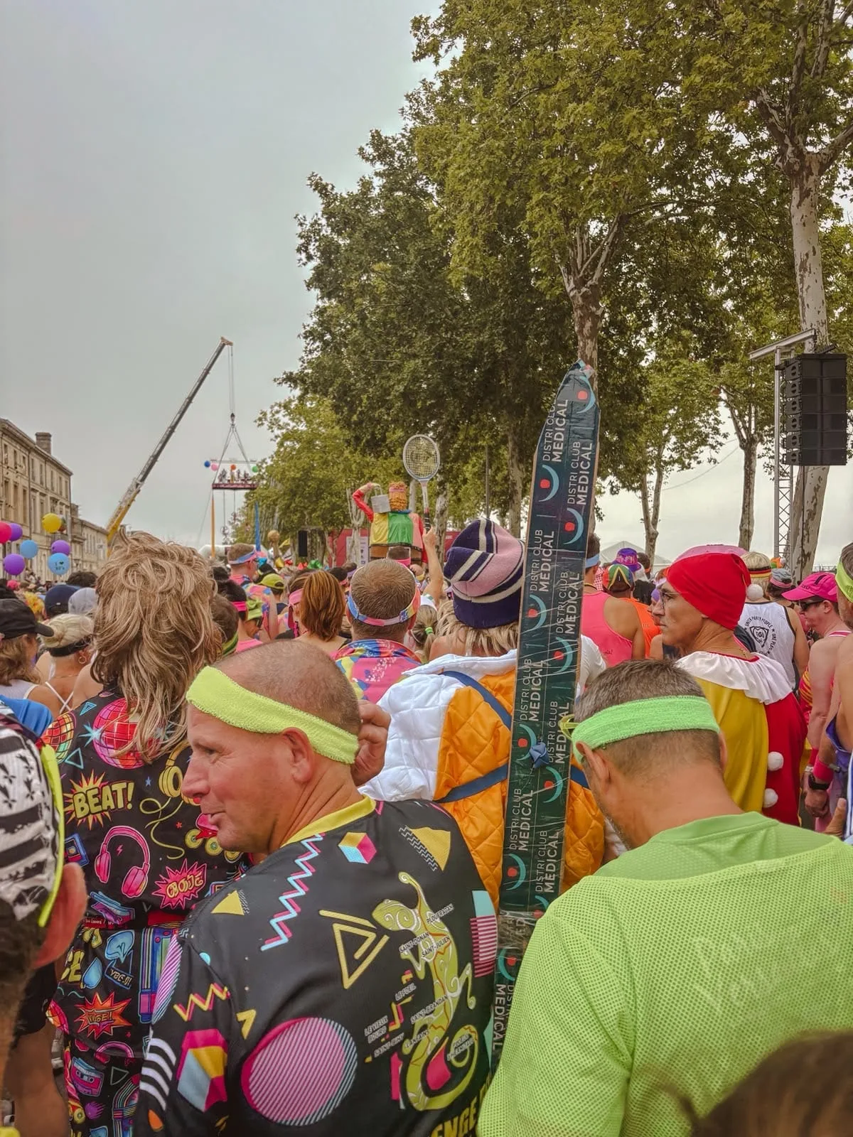 A dense crowd of runners at the start in 1980s costume: neon headbands, a shirt printed with cassettes and disco balls, a giant blonde wig, and one runner carrying a water ski over his shoulder