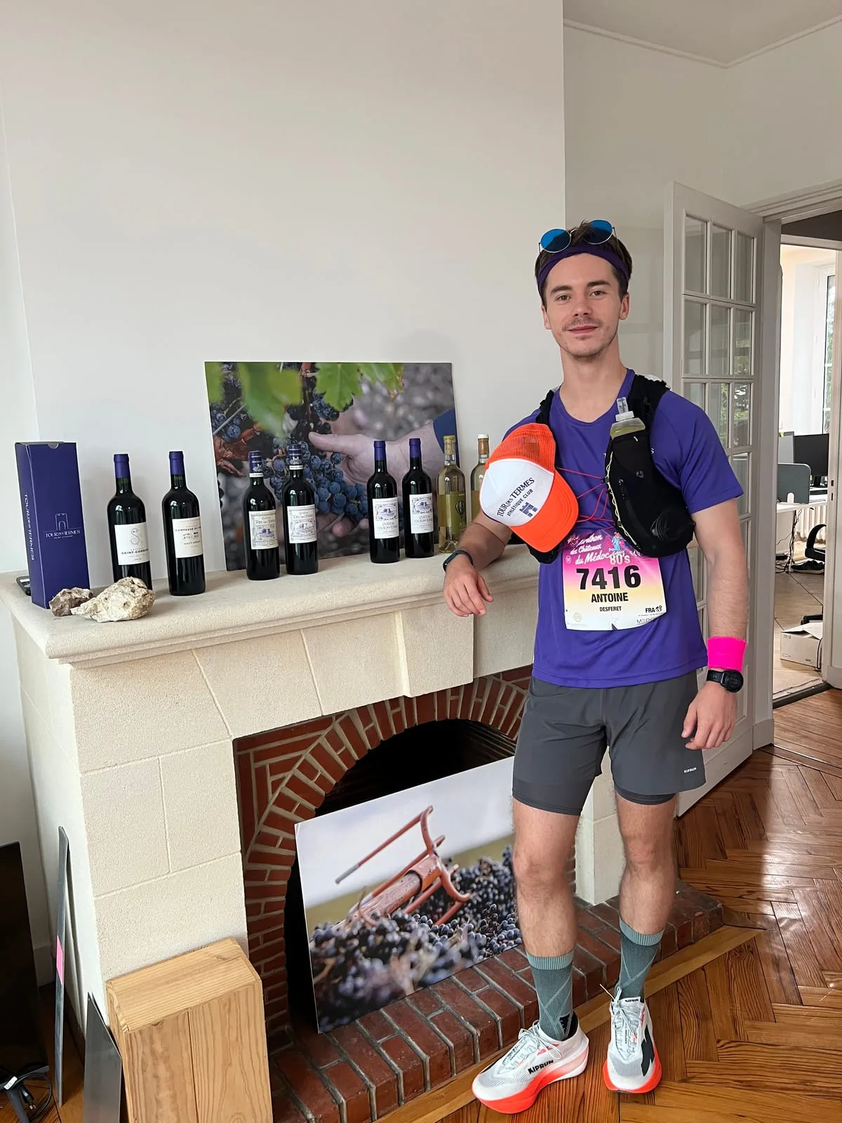 Antoine in a purple Tour des Termes running vest with bib 7416, a purple headband and an orange club cap, standing next to a mantelpiece lined with Château Tour des Termes bottles