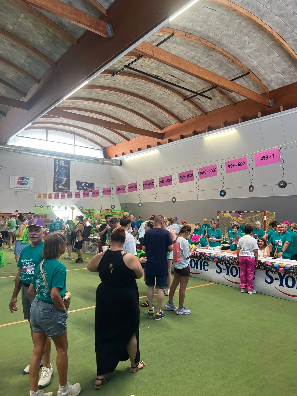 Runners collecting their bibs the day before the race in a sports hall, at a long row of desks staffed by volunteers in green marathon t-shirts, with pink signs overhead marking each bib number range