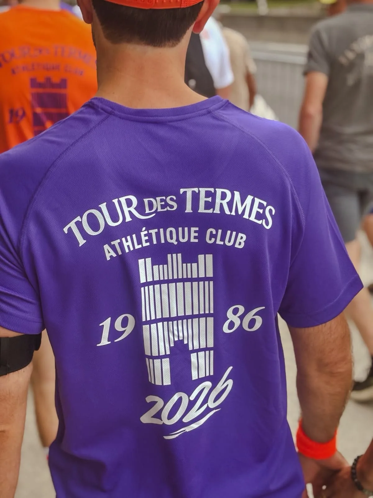 The back of a purple Tour des Termes Athlétique Club running shirt in the crowd at the start, printed with the club's tower logo and the years 1986 and 2026