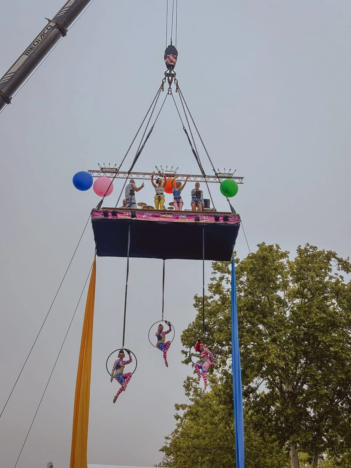 A stage suspended from a crane above the start line, with a band and dancers in 80s outfits waving from the platform and three aerialists performing on hoops below it