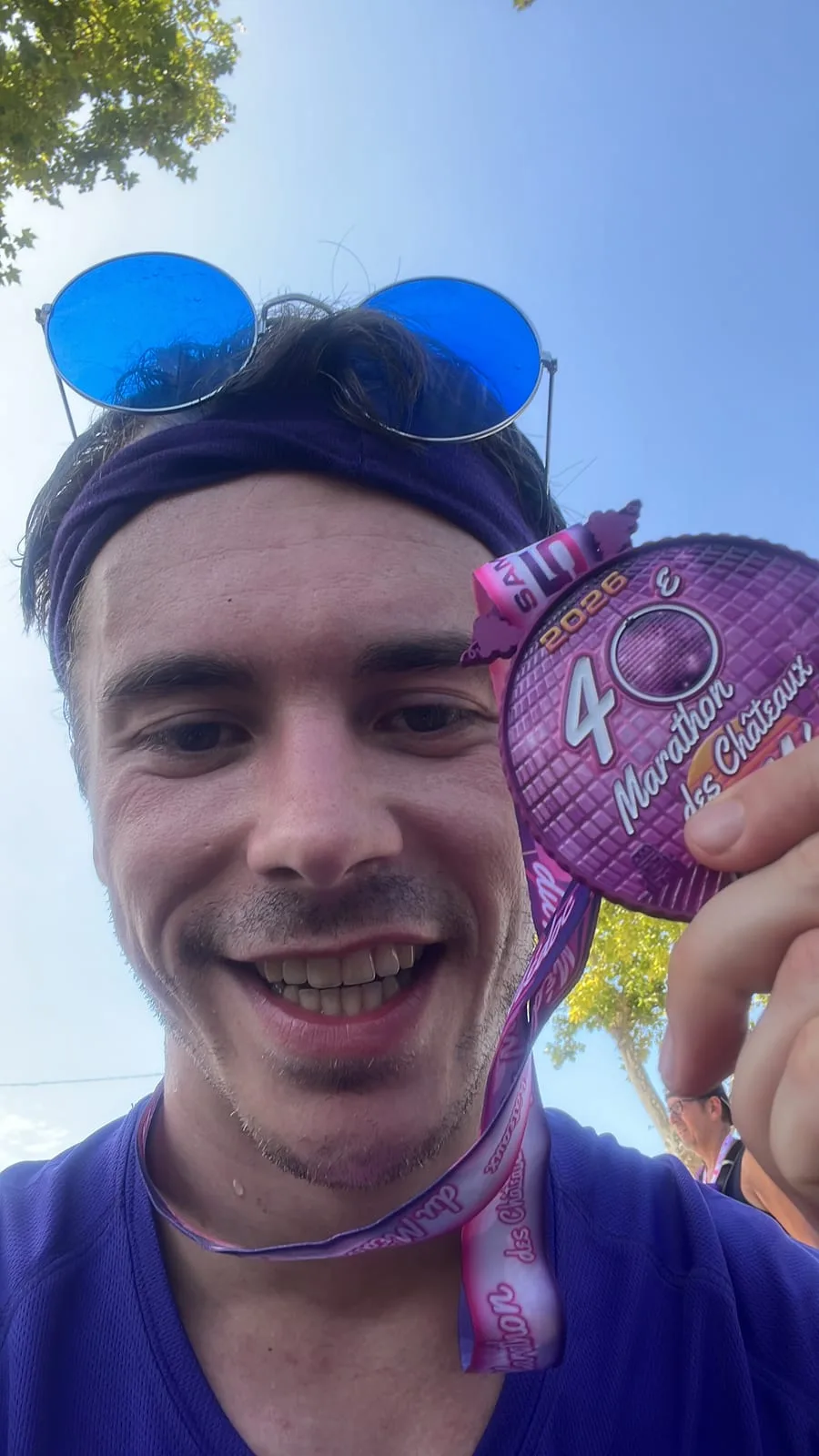 A finish-line selfie: Antoine smiling in a purple vest and headband with round blue sunglasses pushed up, holding the pink 40th Marathon des Châteaux du Médoc finisher medal on its ribbon