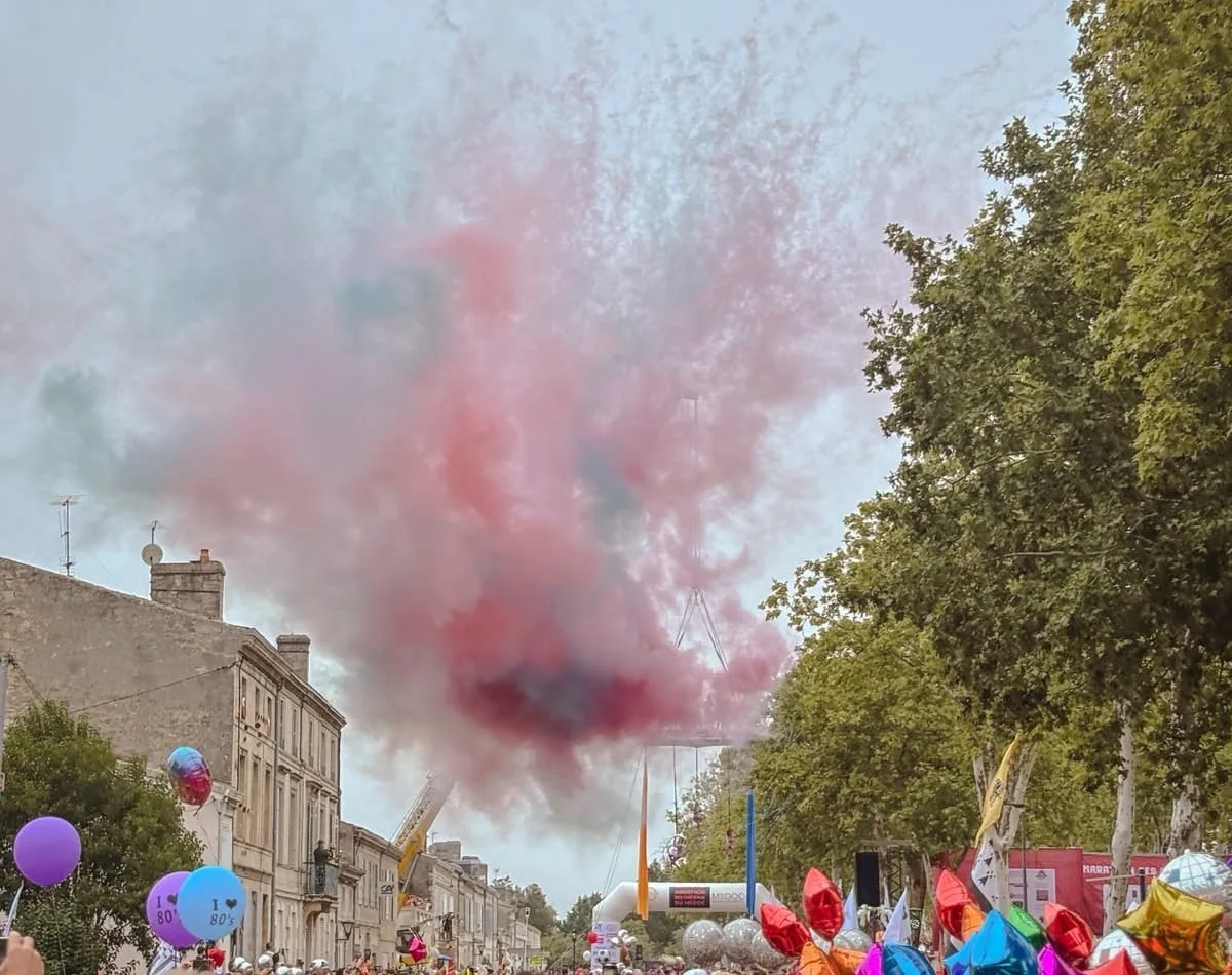 A cloud of pink smoke drifting over the start line of the Marathon du Médoc in Pauillac, with I love 80's balloons and foil stars above a crowd of costumed runners