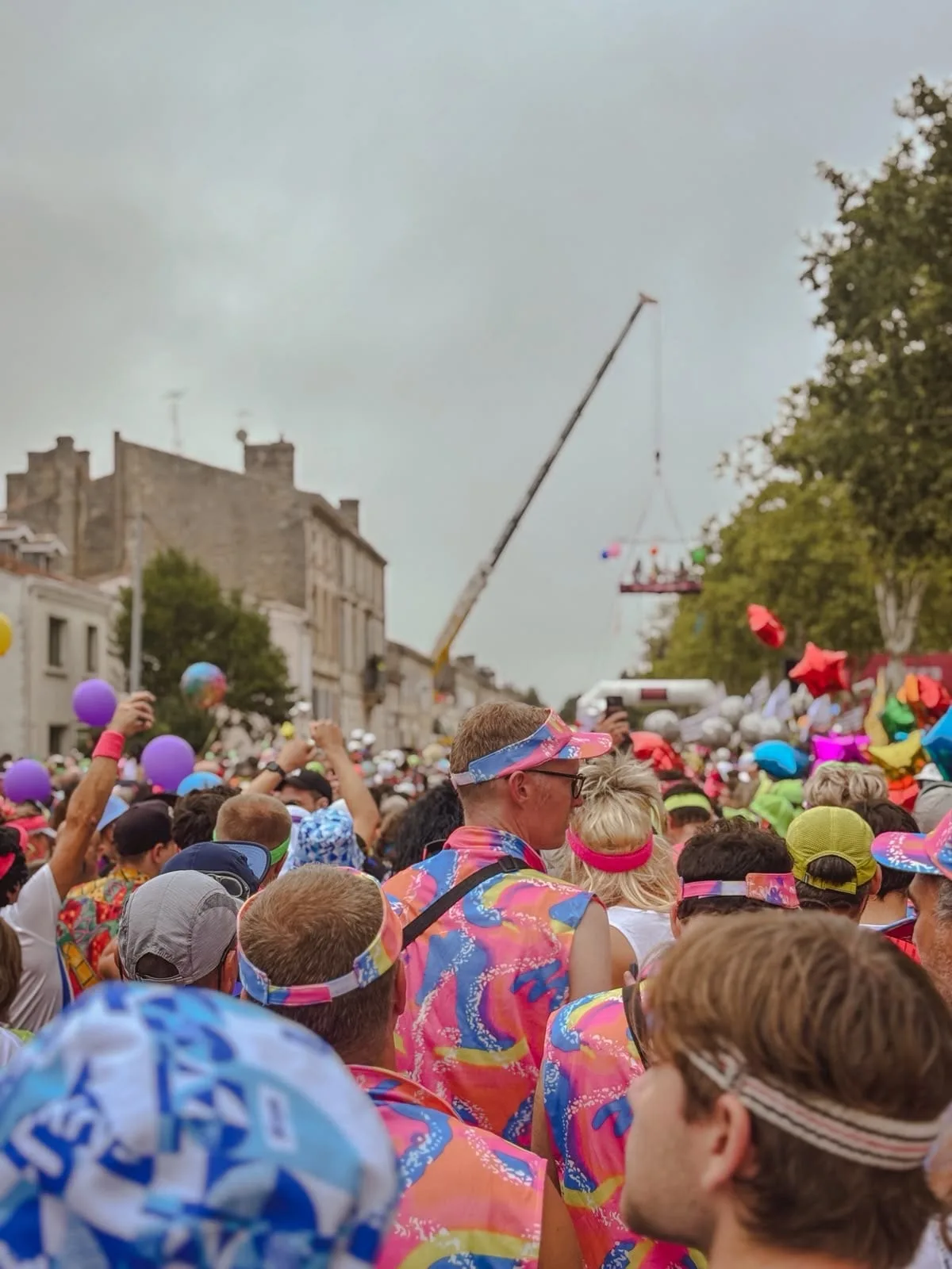 The start pen packed with runners in 1980s neon costumes, headbands and visors, with balloons overhead and a crane holding a suspended stage above the street