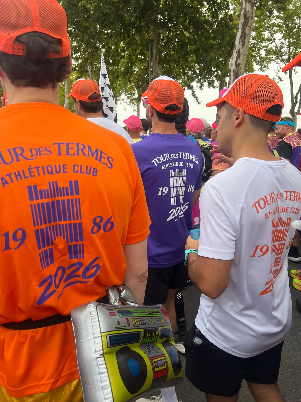 The Tour des Termes team at the start, seen from behind in orange, purple and white club shirts and matching orange caps, one of them carrying an inflatable boombox
