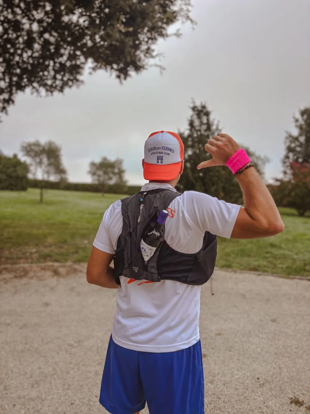 A runner seen from behind on a gravel path through the vines, wearing an orange Tour des Termes cap and a hydration vest with a bottle of red wine in the back pocket, pointing at it with his thumb