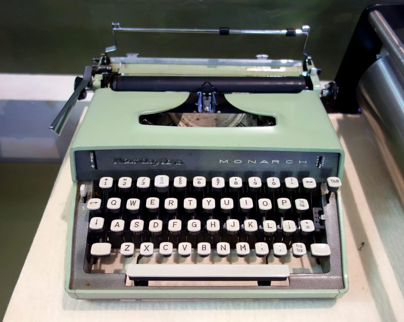 A mint green Remington Monarch portable typewriter with cream keys, displayed on a white plinth in a museum case