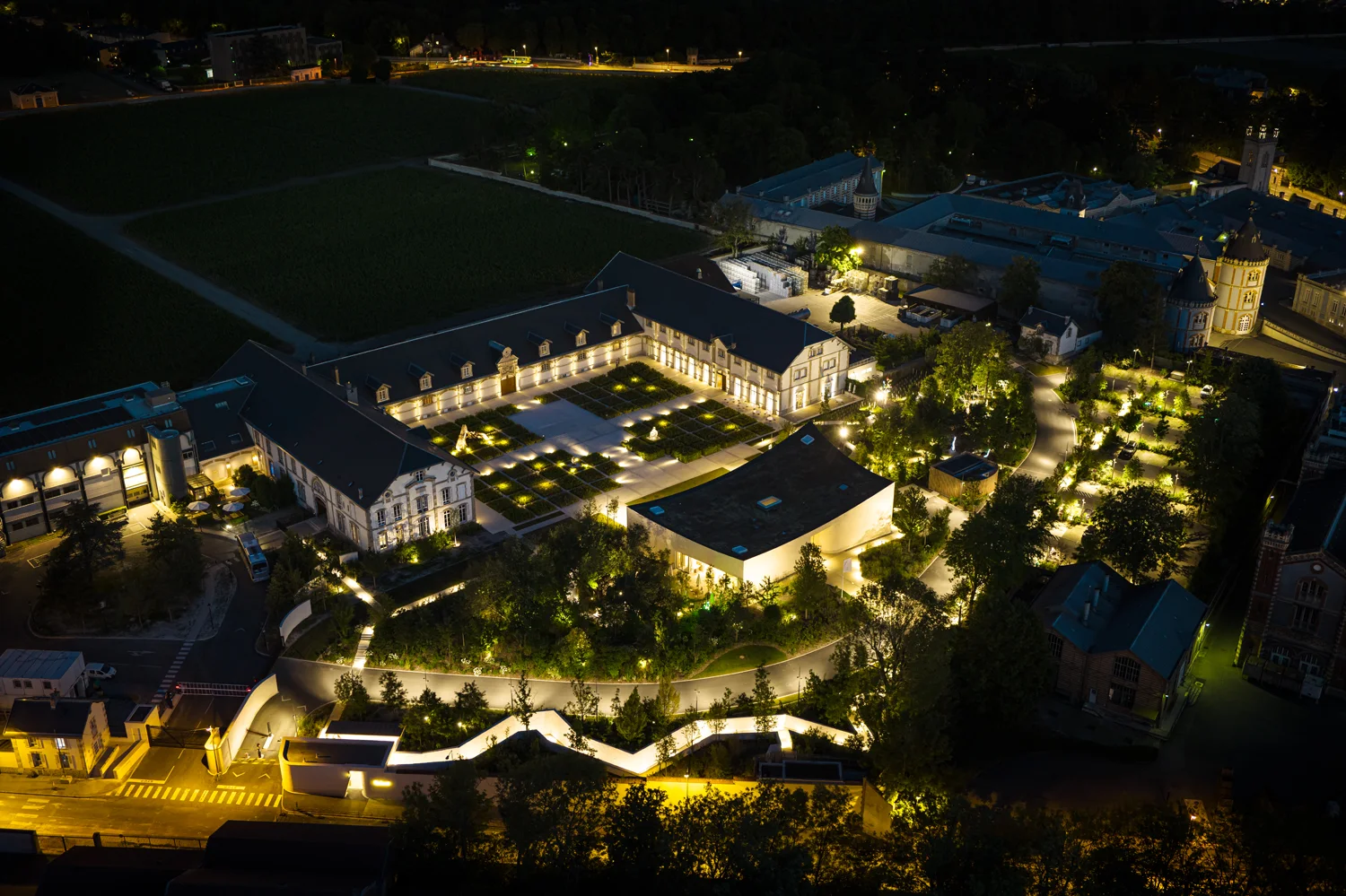 Aerial night view of Ruinart's 4 rue des Crayères in Reims, the historic courtyard and gardens lit from below, with vineyard rows visible at the edge of the city