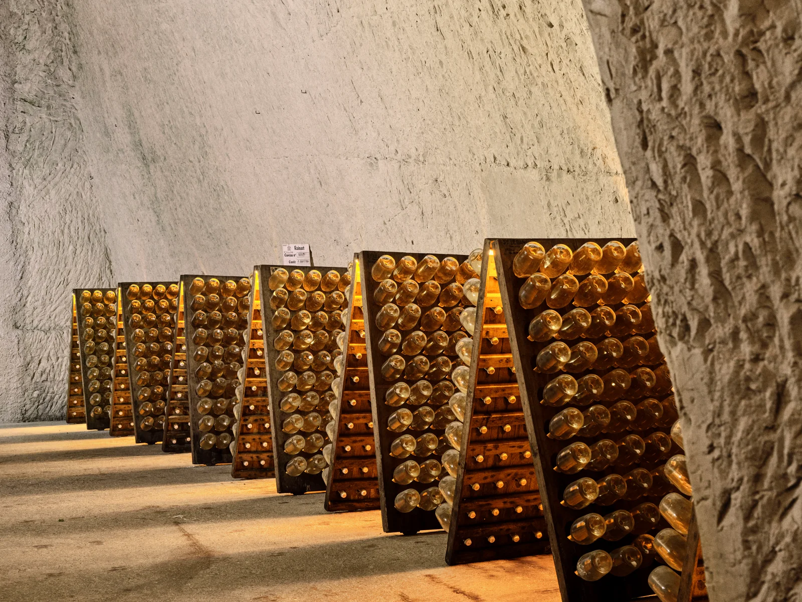 Rows of wooden riddling racks holding Champagne bottles, lit from within, against the rough chalk wall of a Ruinart crayère