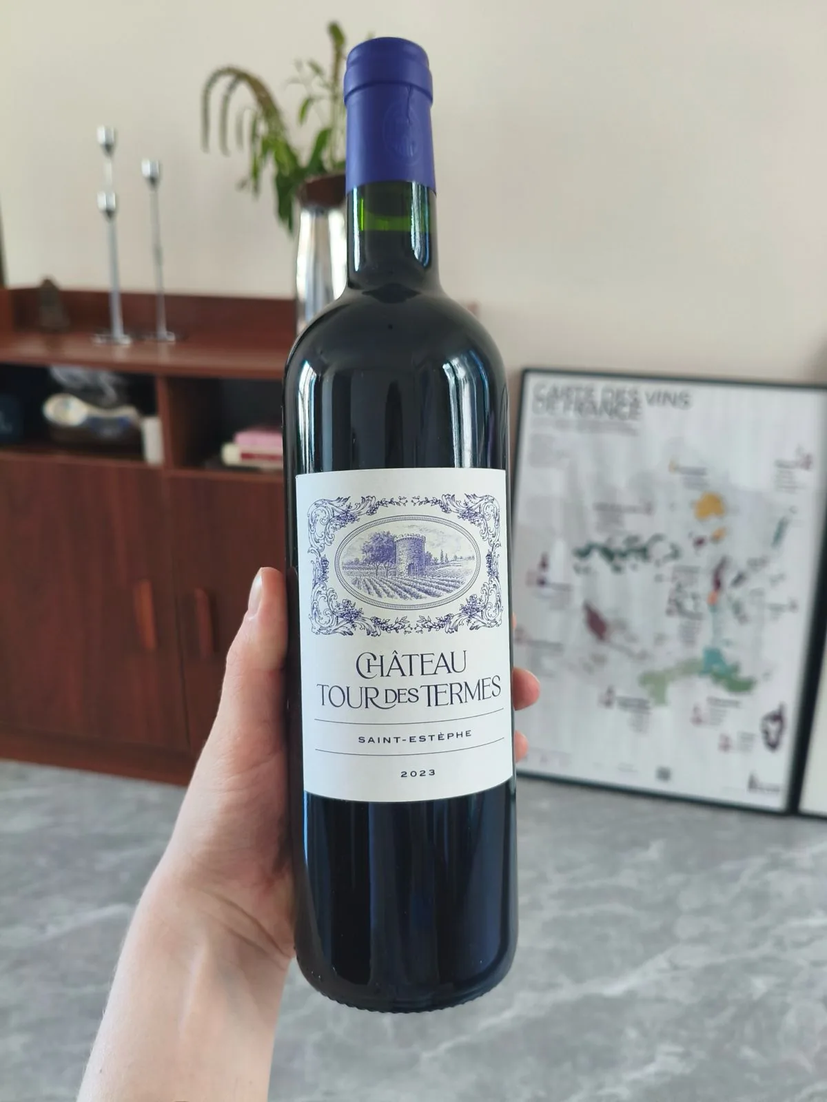 The full bottle of Château Tour des Termes 2023 with its purple capsule, held up in a living room in front of a framed French wine map