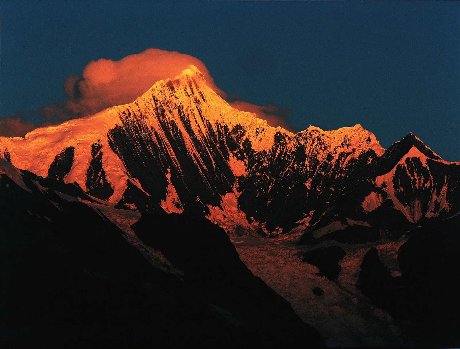 The summit of Kawagebo in the Meili Snow Mountains, Deqin, Yunnan, lit orange at sunrise above dark valleys, close to where Xige is building its Yunnan winery