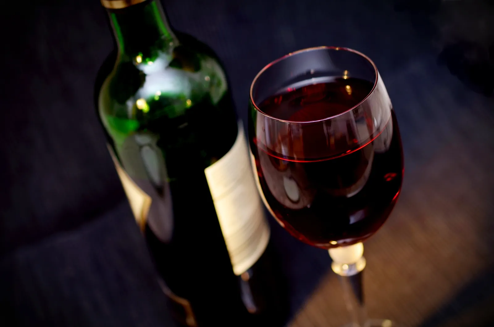 A bottle of red wine with a plain label next to a poured glass of red, on a dark background