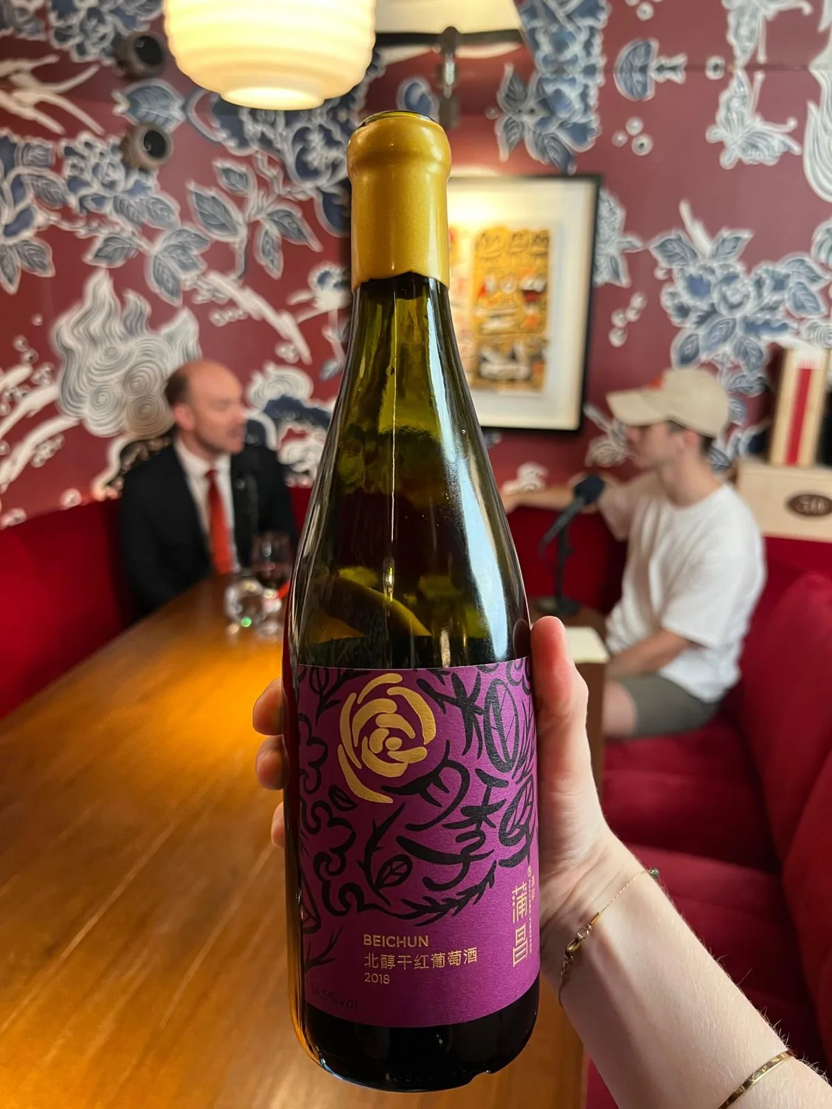 The bottle of Puchang Beichun 2018 held up in a room with deep red walls and large blue and white floral wallpaper, a recording table out of focus behind it