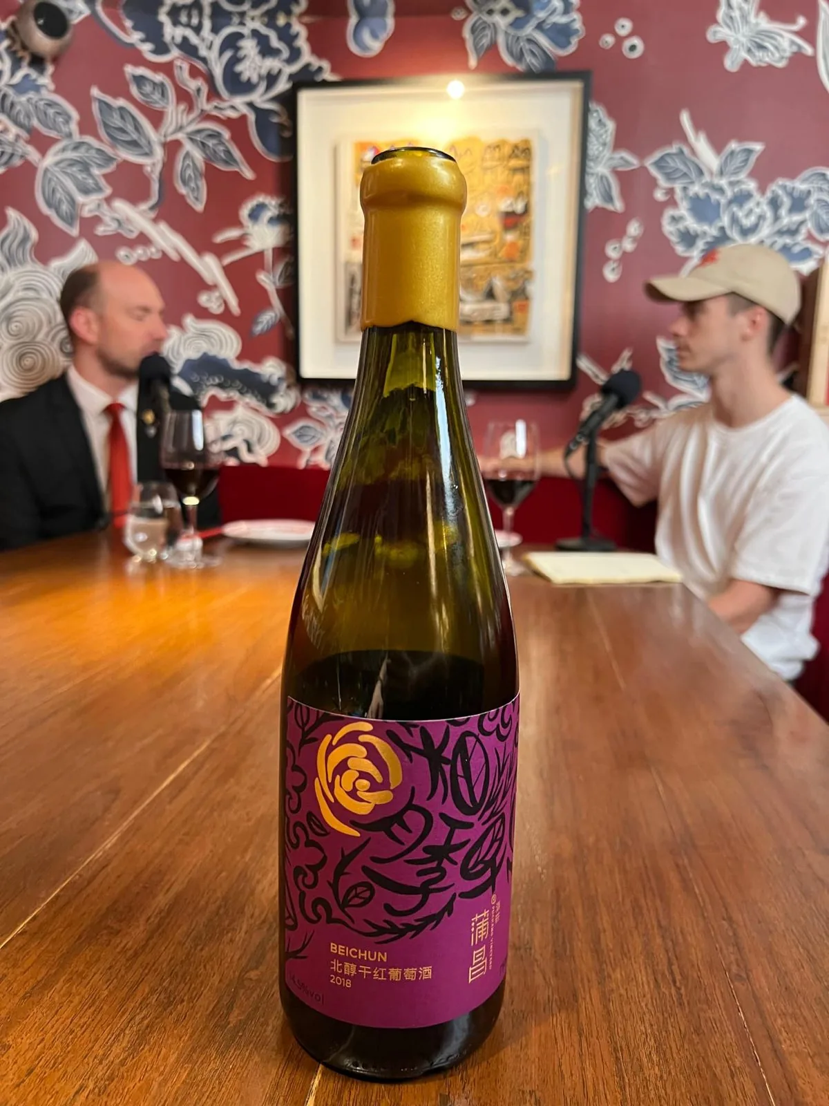 The bottle of Puchang Beichun 2018 with its gold wax-style capsule standing on a wooden table during a podcast recording, two people out of focus at their microphones behind it and glasses of red wine on the table